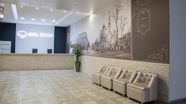 Et hotellets reception område med et moderne design, der inkluderer en receptionsdisk, et siddeområde med sofaer, et stort muralmaleri på væggen og en potteplante.