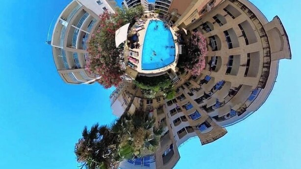 Vista aérea de un hotel con piscina central, rodeado de balcones y un cielo azul despejado.