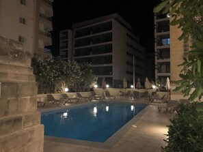 Un área de piscina nocturna con sillas de descanso y sombrillas, rodeada de edificios.