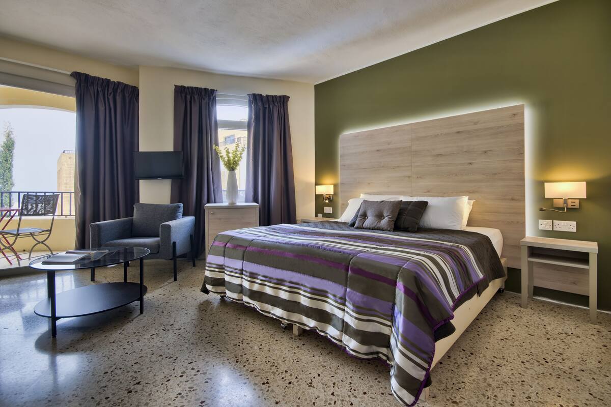 Habitación de hotel con cama, una silla, una mesita y un balcón con cortinas.