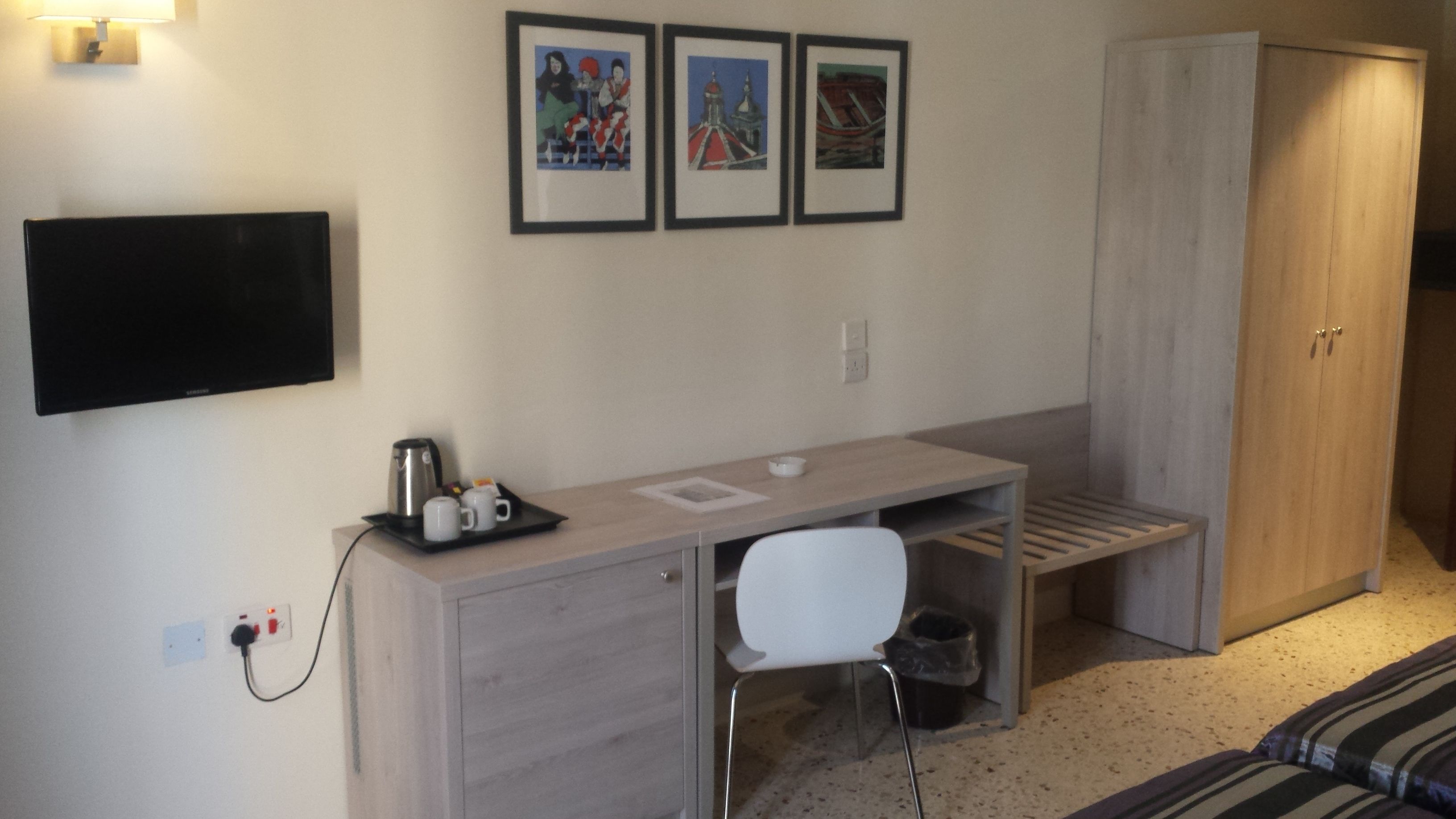 A hotel room with a desk, chair, and a wardrobe. There are three framed pictures on the wall.