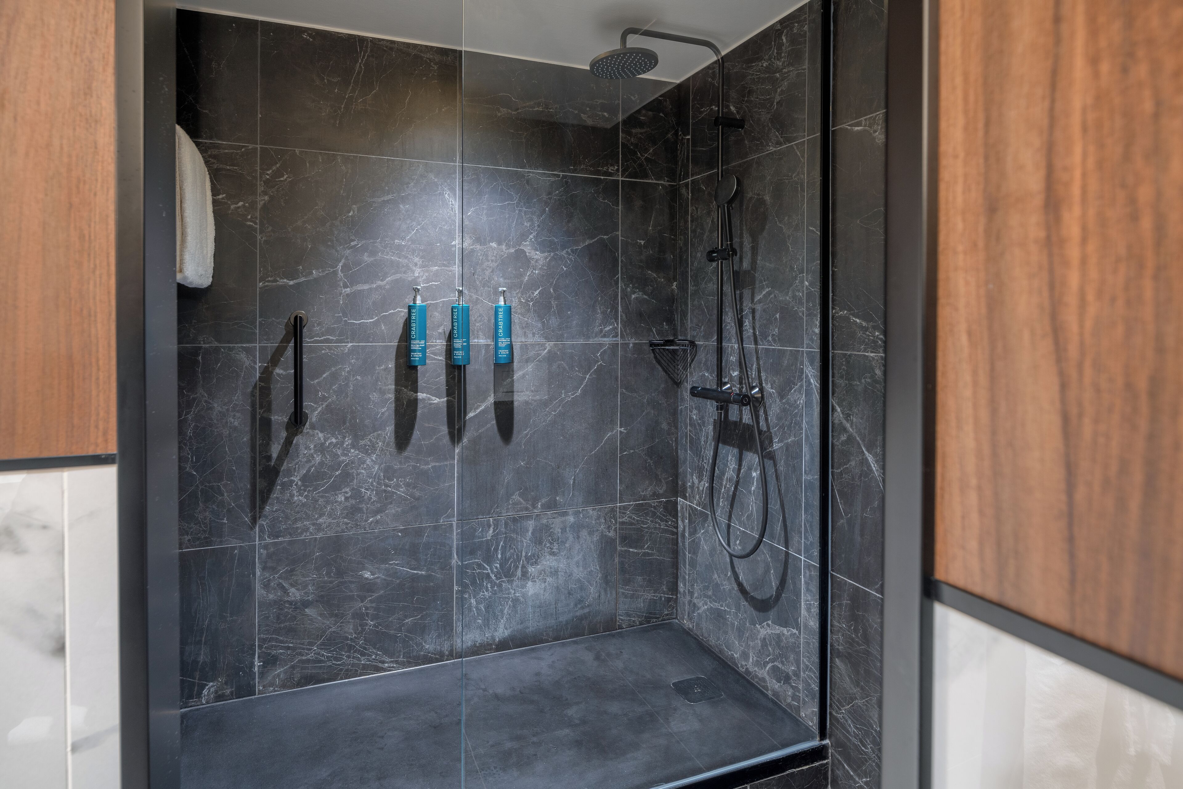 A modern shower with dark marble tiles, a handheld showerhead, and two bottles of shampoo.