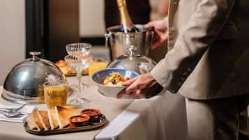 A hotel room with a dining table set for a meal, a bottle of champagne, and a person in a suit standing by the table.