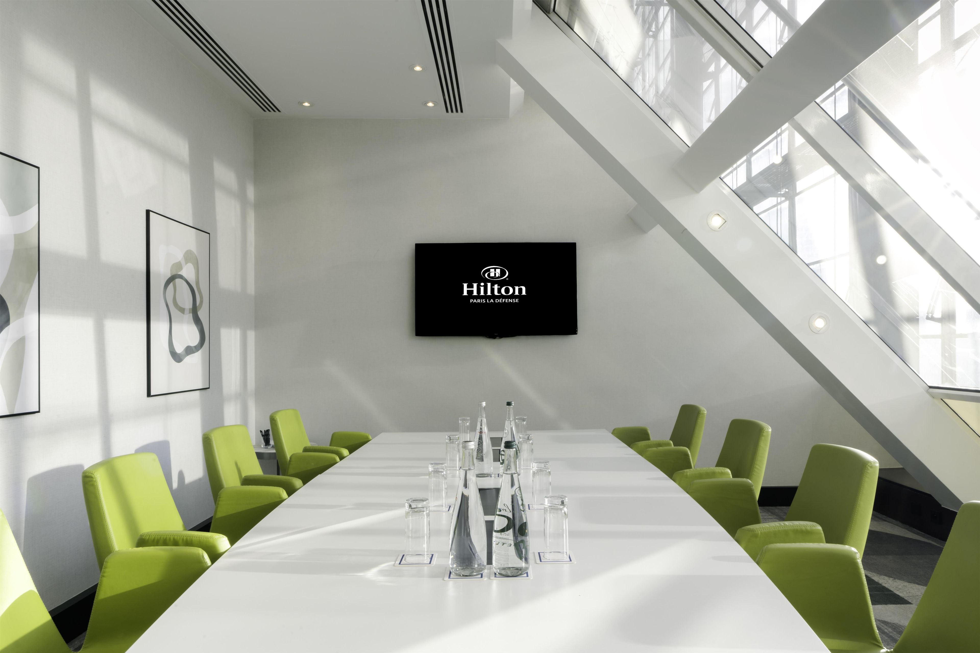 A conference room with a long table, green chairs, a flat-screen TV, and a large window.