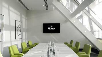 A conference room with a long table, green chairs, a flat-screen TV, and a large window.