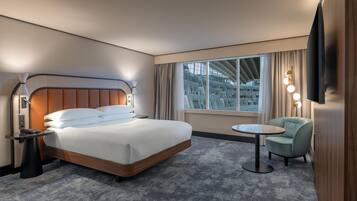 Ett hotellrum med en stor säng, ett nattduksbord, en lampa, en stol och utsikt över en byggnad genom fönstret.