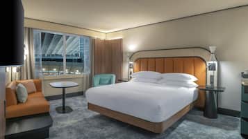 A hotel room with a large bed, a sitting area with a sofa and armchair, a small round table, and a view of a building through large windows.