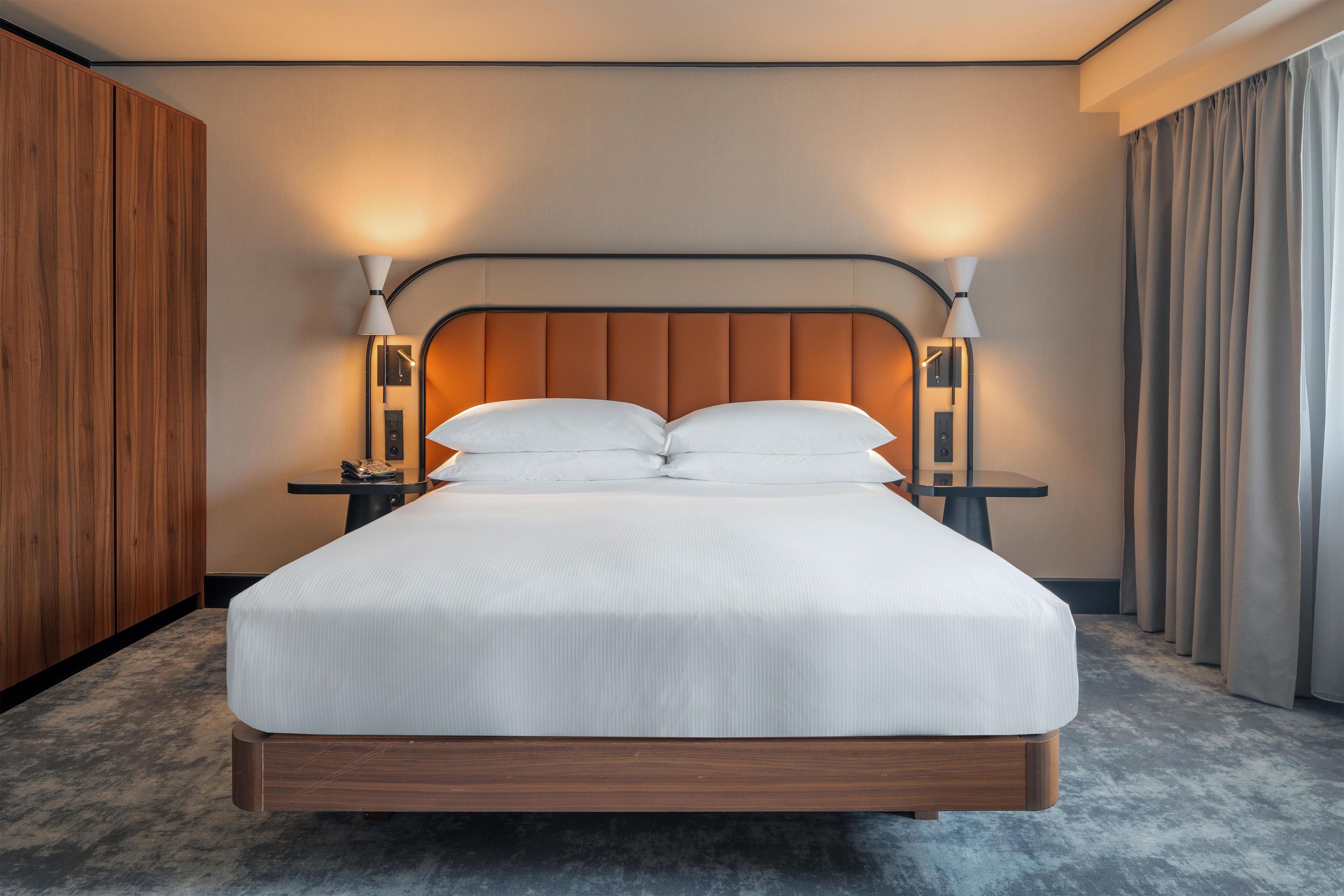A hotel room with a large bed, two bedside tables, a wooden headboard, and a nightstand with a lamp.
