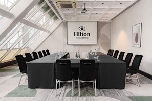Un salón de conferencias con una larga mesa, sillas negras y una pantalla de proyector que muestra el logotipo de Hilton.