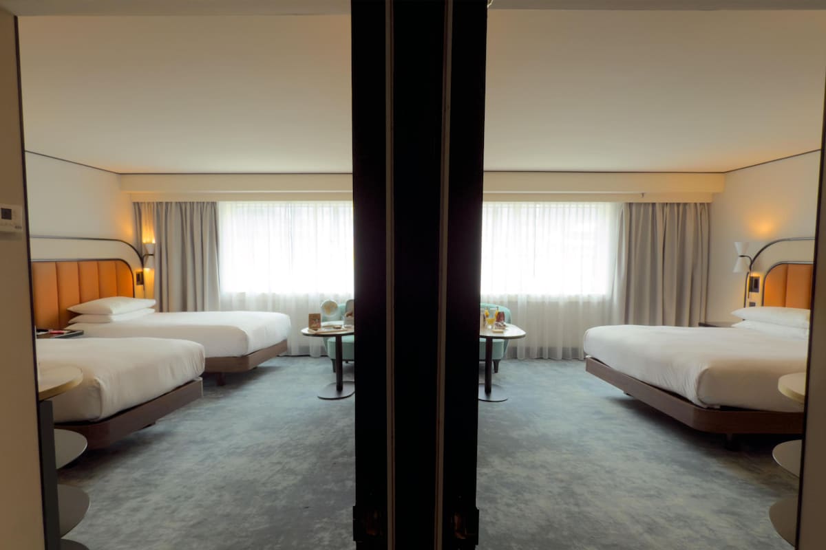 A hotel room with two beds, a small table, and a large window with curtains.