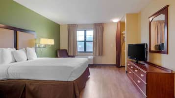 A hotel room with a large bed, a television on a wooden dresser, a window with curtains, and a wall-mounted lamp.