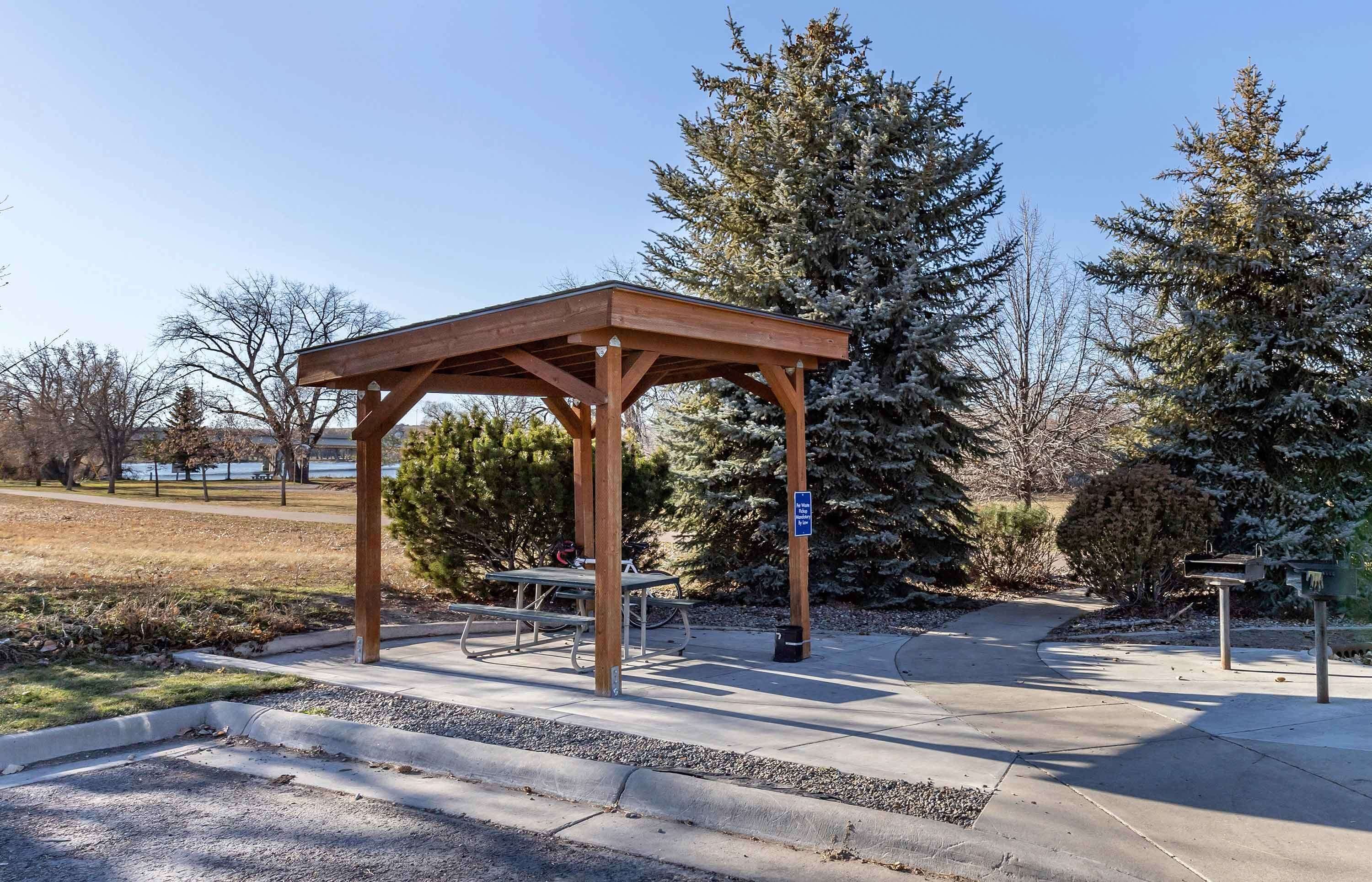 Un kiosque en bois avec des bancs, une boîte aux lettres et un panneau indicateur, situé dans un parc où les arbres sont recouverts de neige.