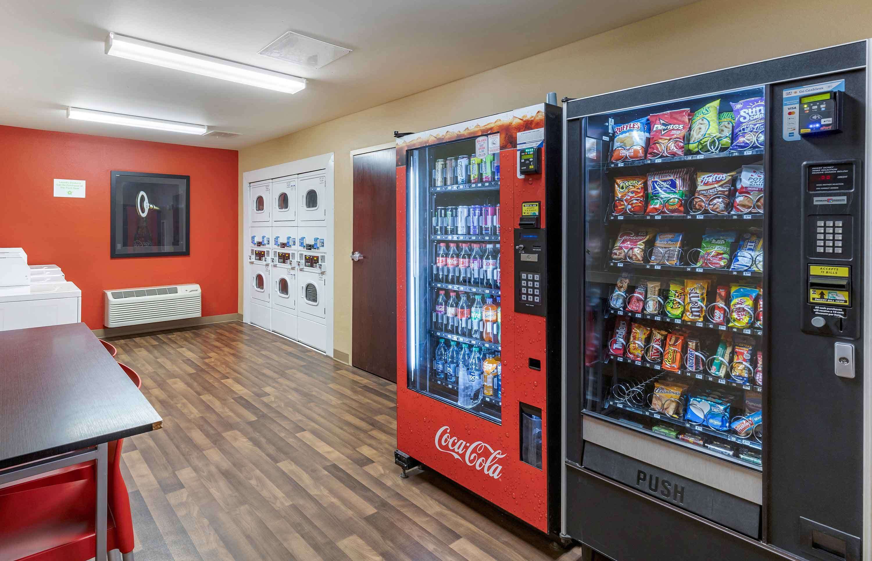 A room with a Coca-Cola vending machine, a snack vending machine, a table, and a microwave.