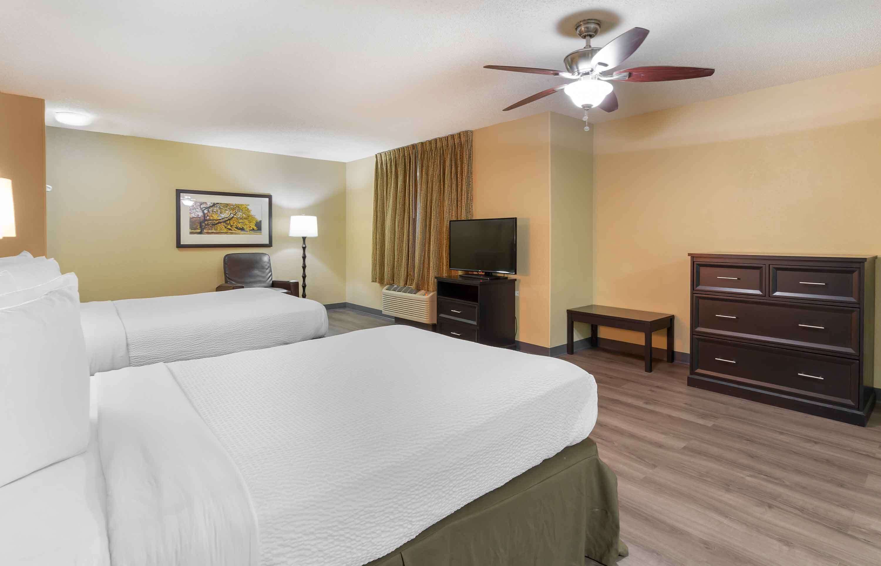 A hotel room with two beds, a television, a desk, a chair, a wardrobe, and a ceiling fan.