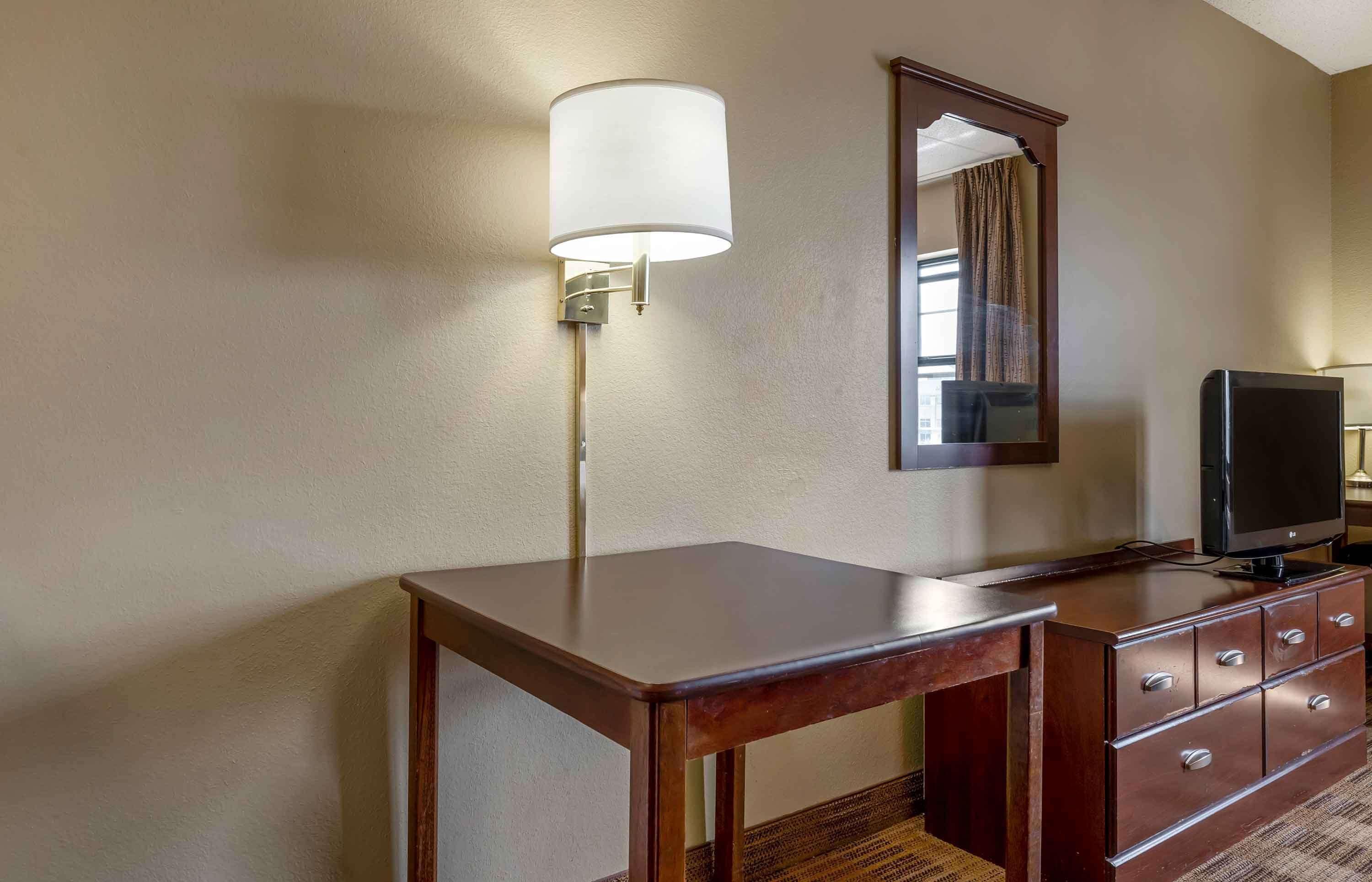 A hotel room with a wooden desk, a television on a dresser, a wall-mounted lamp, and a mirror.