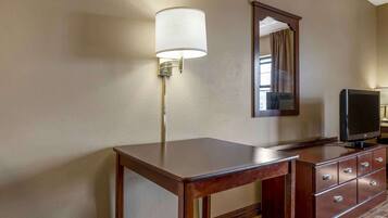 A hotel room with a wooden desk, a television on a dresser, a wall-mounted lamp, and a mirror.