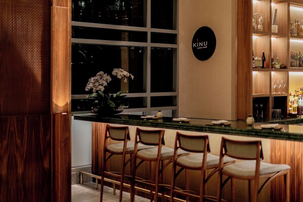 Zona de bar con sillas de madera, letrero de barra de sushi y una ventana con orquídeas.