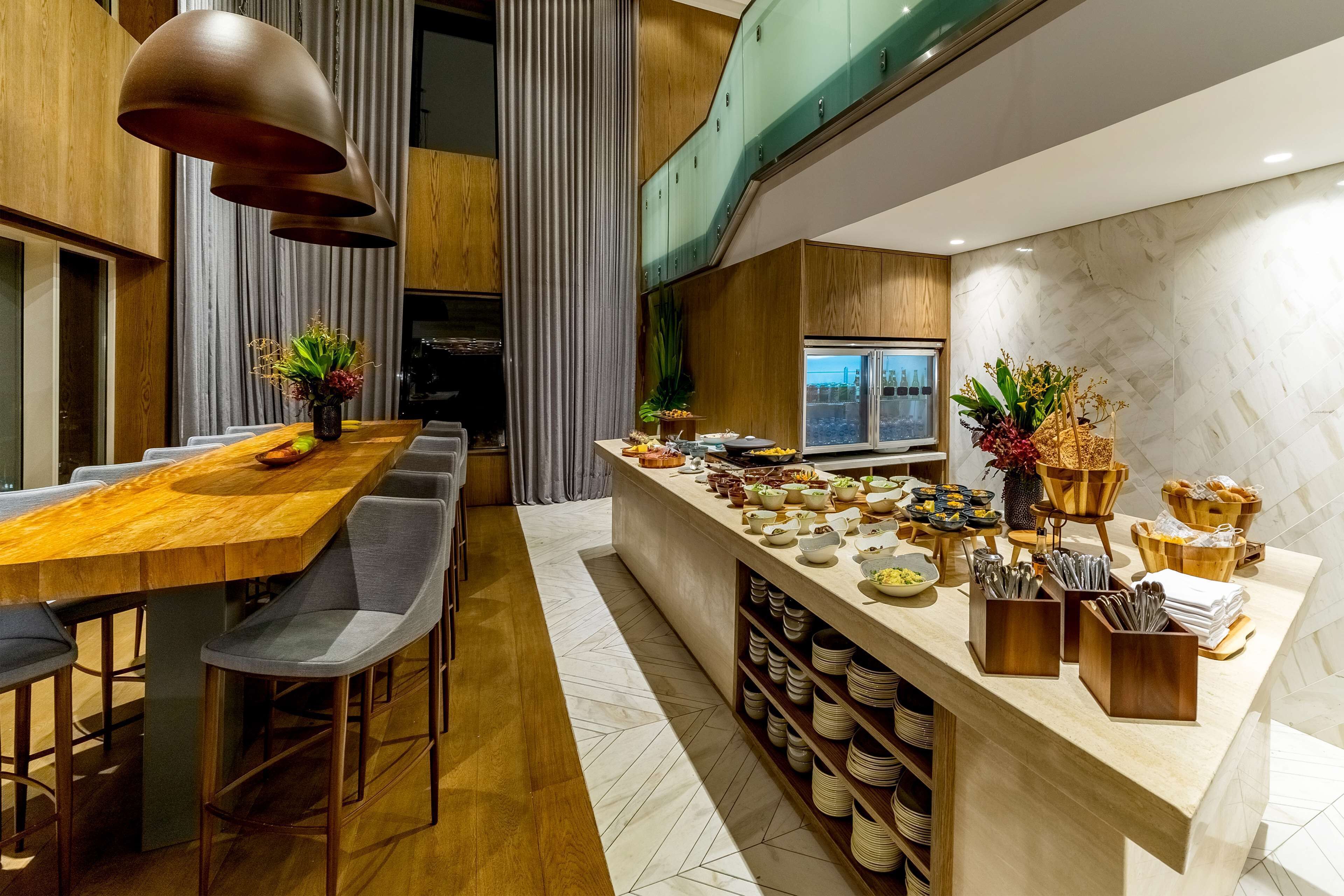 A modern dining area with a wooden table and grey chairs, a buffet counter with various food items, and a view of the city through a window.