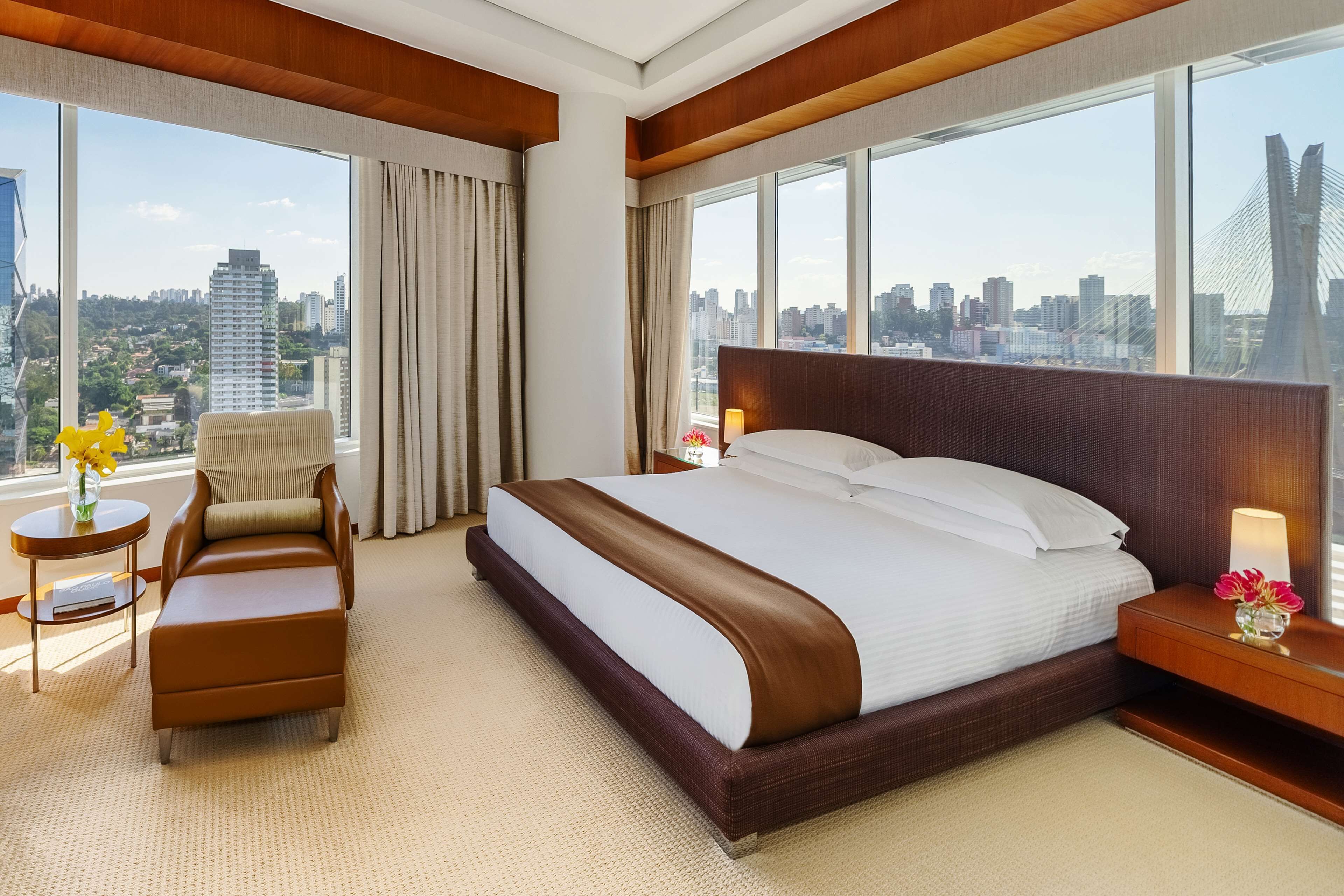 Quarto de hotel com uma cama grande, uma poltrona, uma mesinha e vista para a paisagem urbana através de amplas janelas.