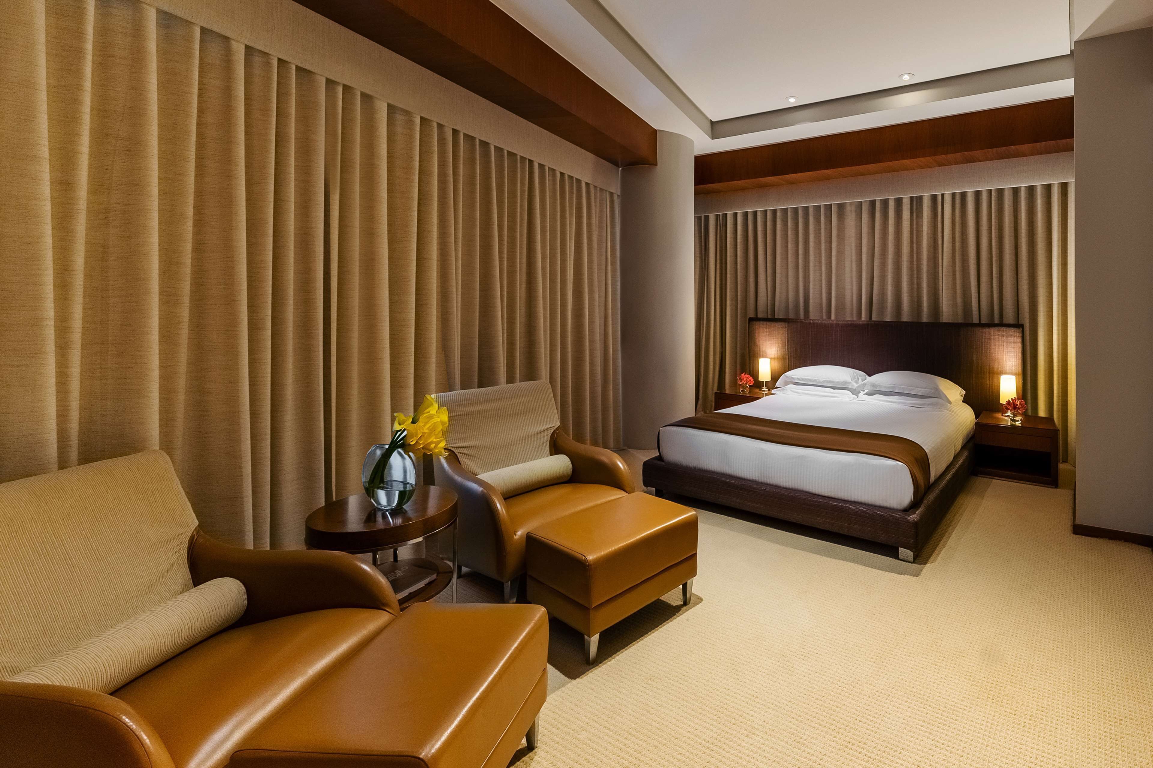 Un cuarto de hotel con una cama grande, un sofá, una silla, una mesita con un florero y una mesita de noche con lámparas.