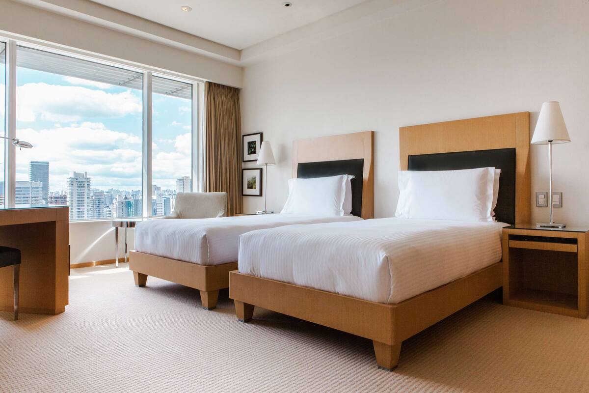 Habitación de hotel con dos camas, un escritorio y un ventanal grande con vistas a la ciudad.