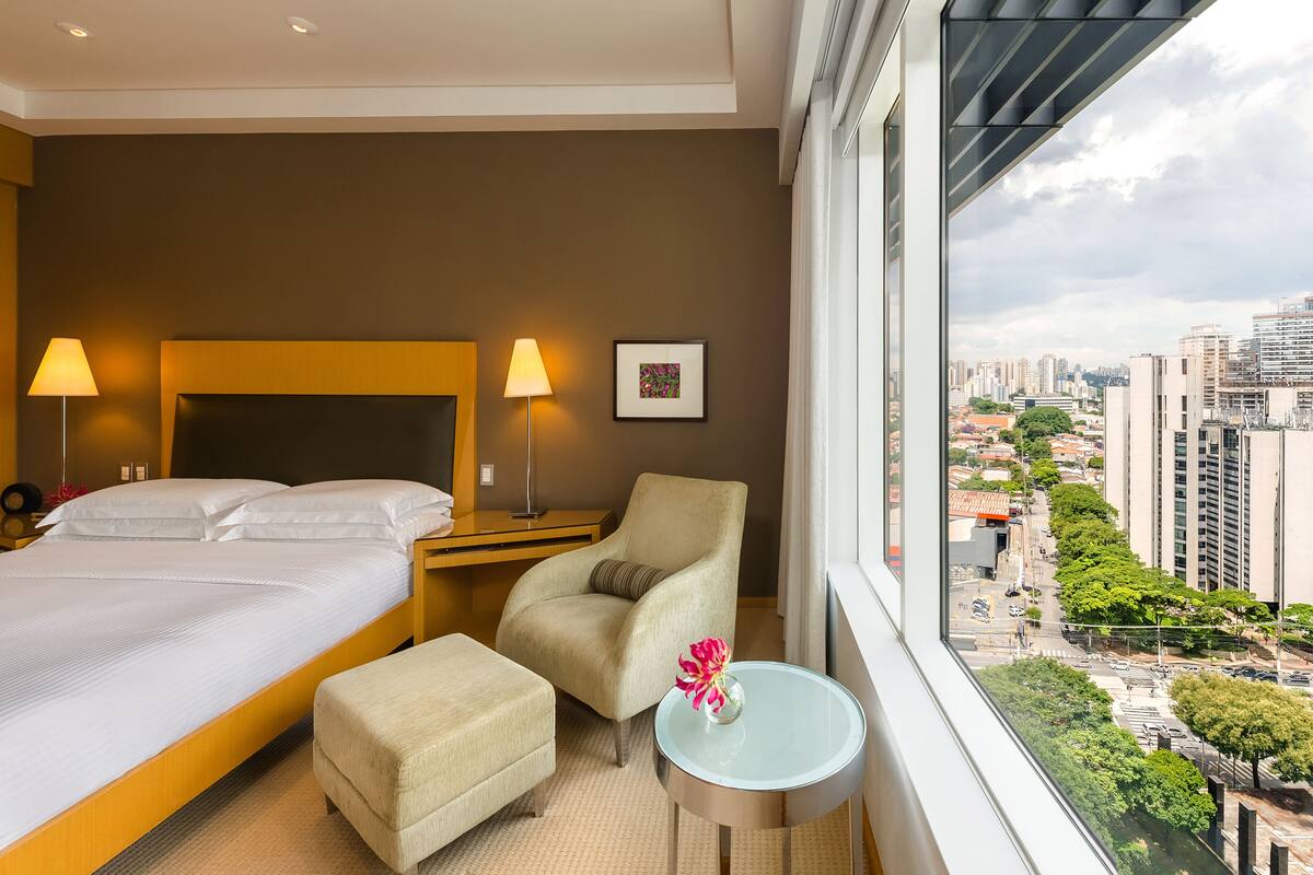 Habitación de hotel con cama, una silla, una mesita con un jarrón y un ventanal con vistas a la ciudad.