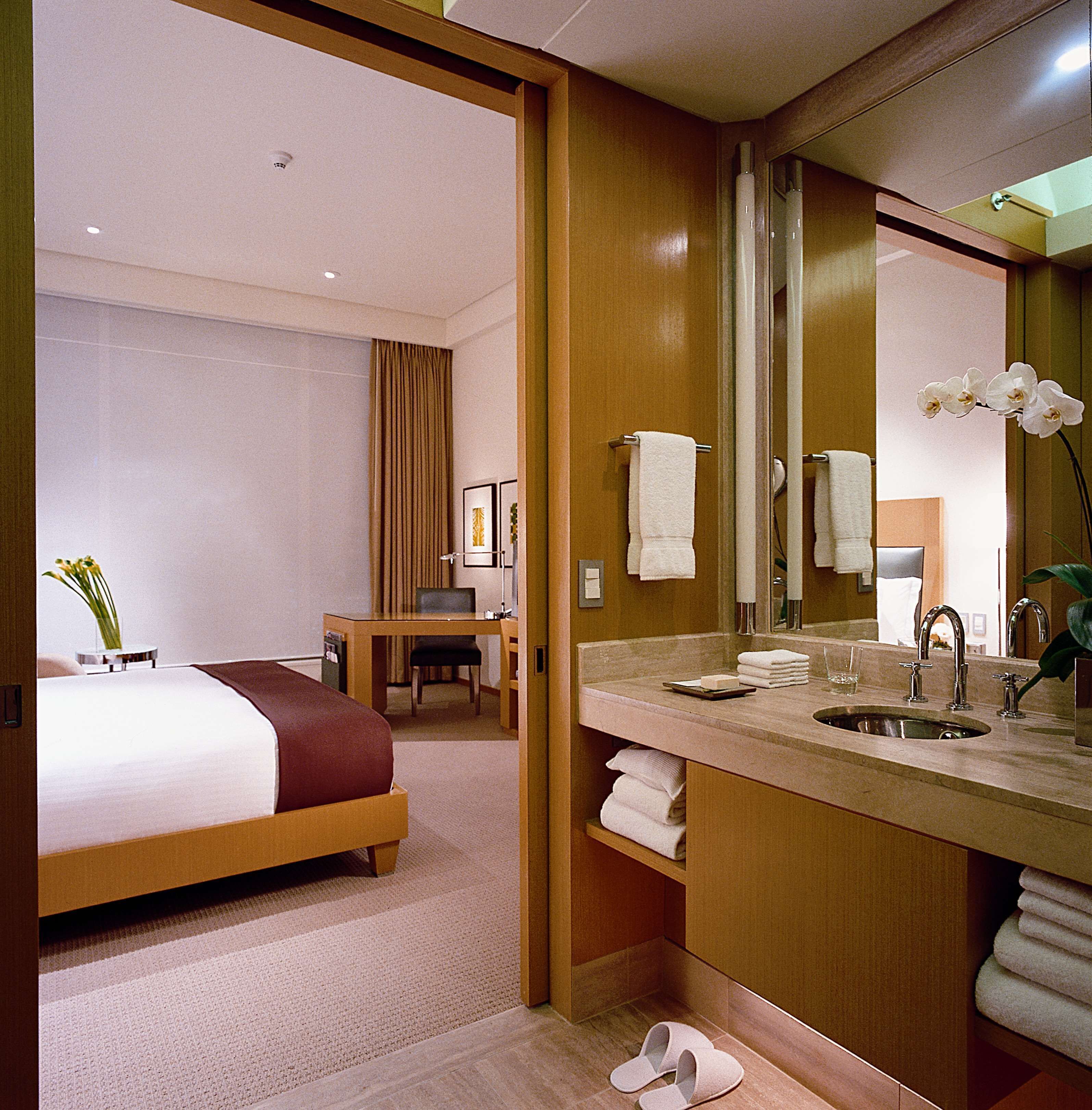 A hotel room with a bed, a desk, a chair, a bathroom with a sink and mirror, and a pair of slippers on the floor.