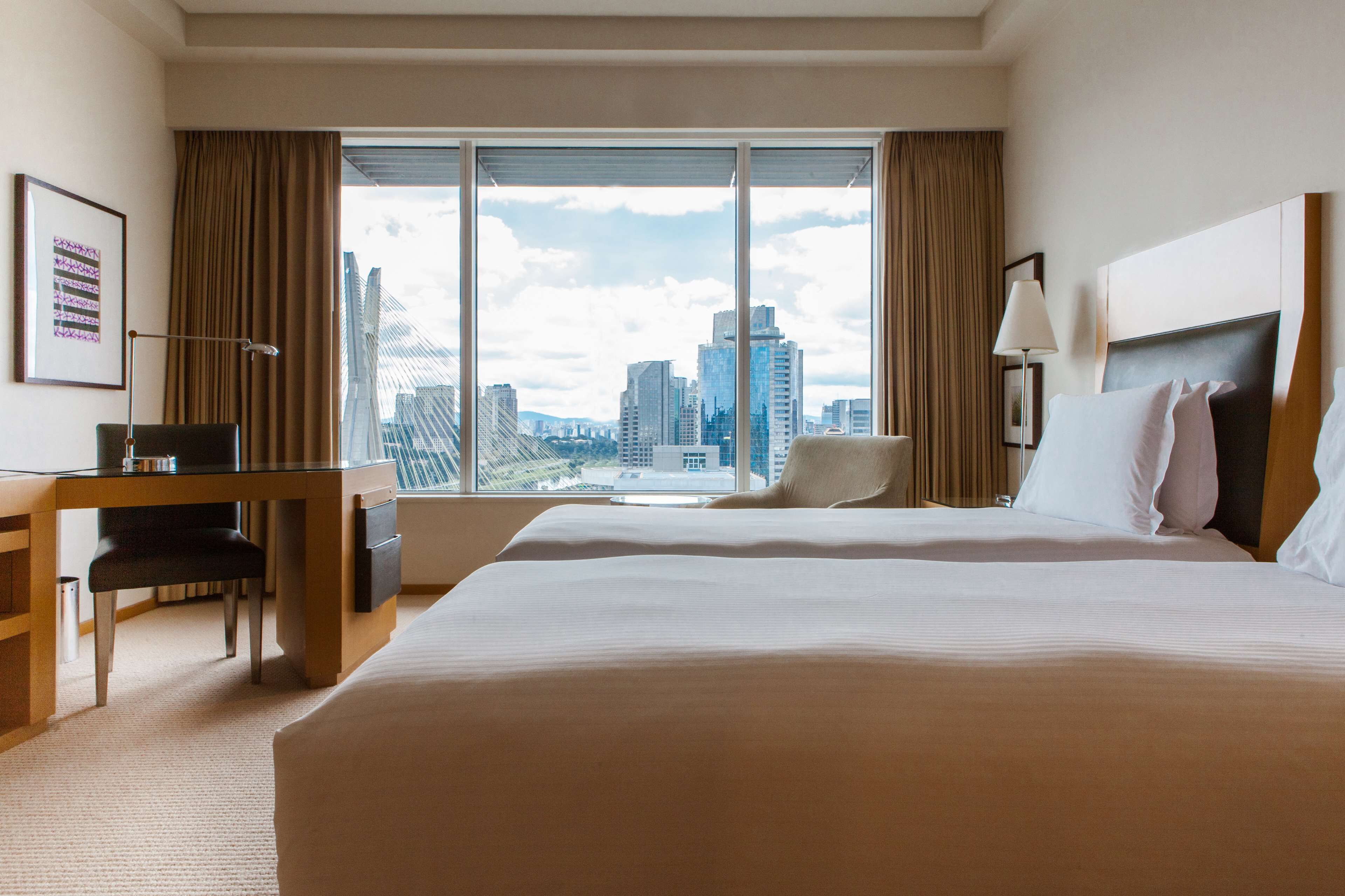 A hotel room with a large bed, a desk, and a chair. There is a large window with a city view.