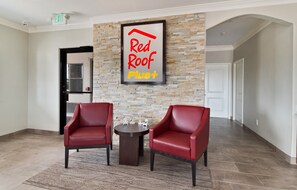 A hallway with two red leather chairs, a small table, and a framed sign that reads 'Red Roof Plus+'. - Red Roof Inn PLUS+ Galveston - Beachfront (Galveston)