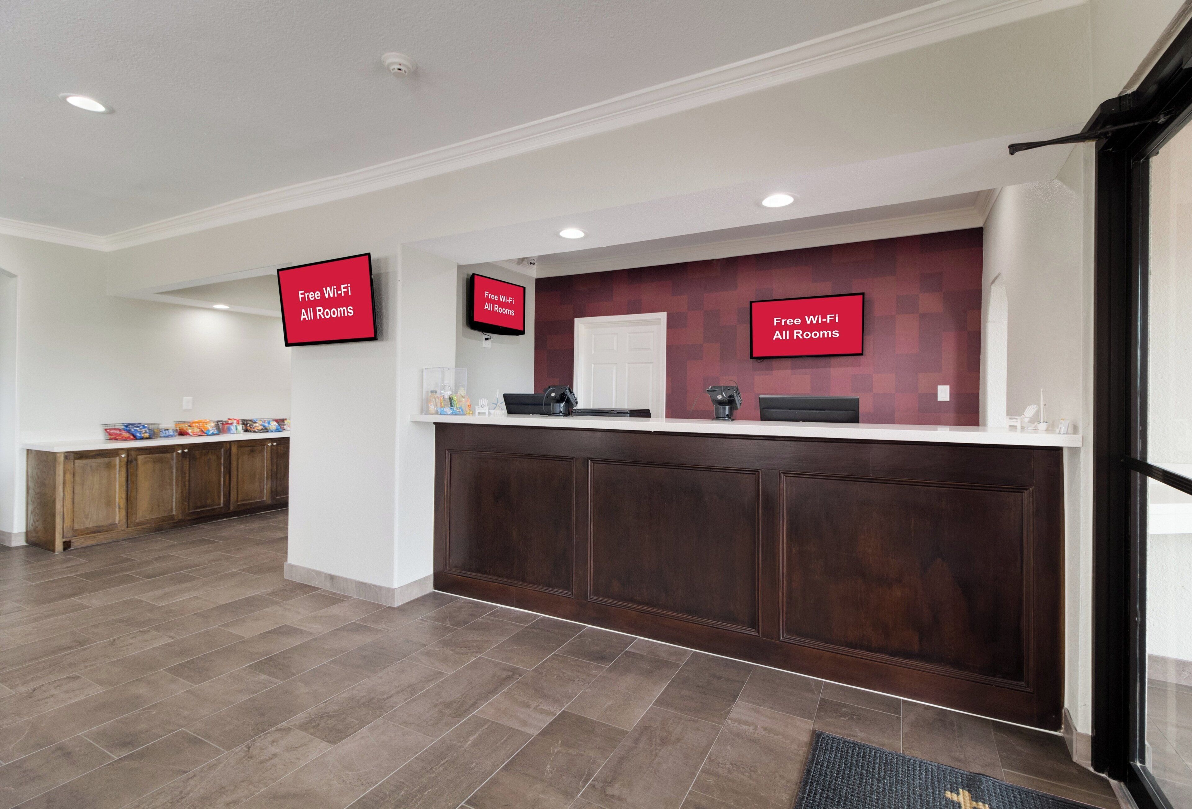 A reception area with a wooden reception desk, a flat-screen TV displaying 'Free Wi-Fi All Rooms,' and a kitchen area with cabinets and a countertop.
