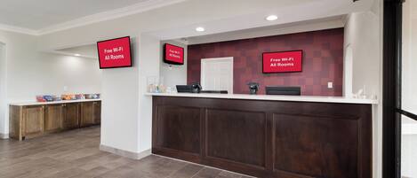 A reception area with a wooden reception desk, a flat-screen TV displaying 'Free Wi-Fi All Rooms,' and a kitchen area with cabinets and a countertop.