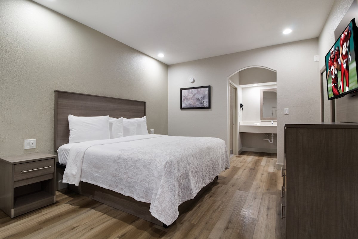 Un dormitorio con una cama grande, una mesita de noche, un aparador, un espejo y un televisor montado en la pared.