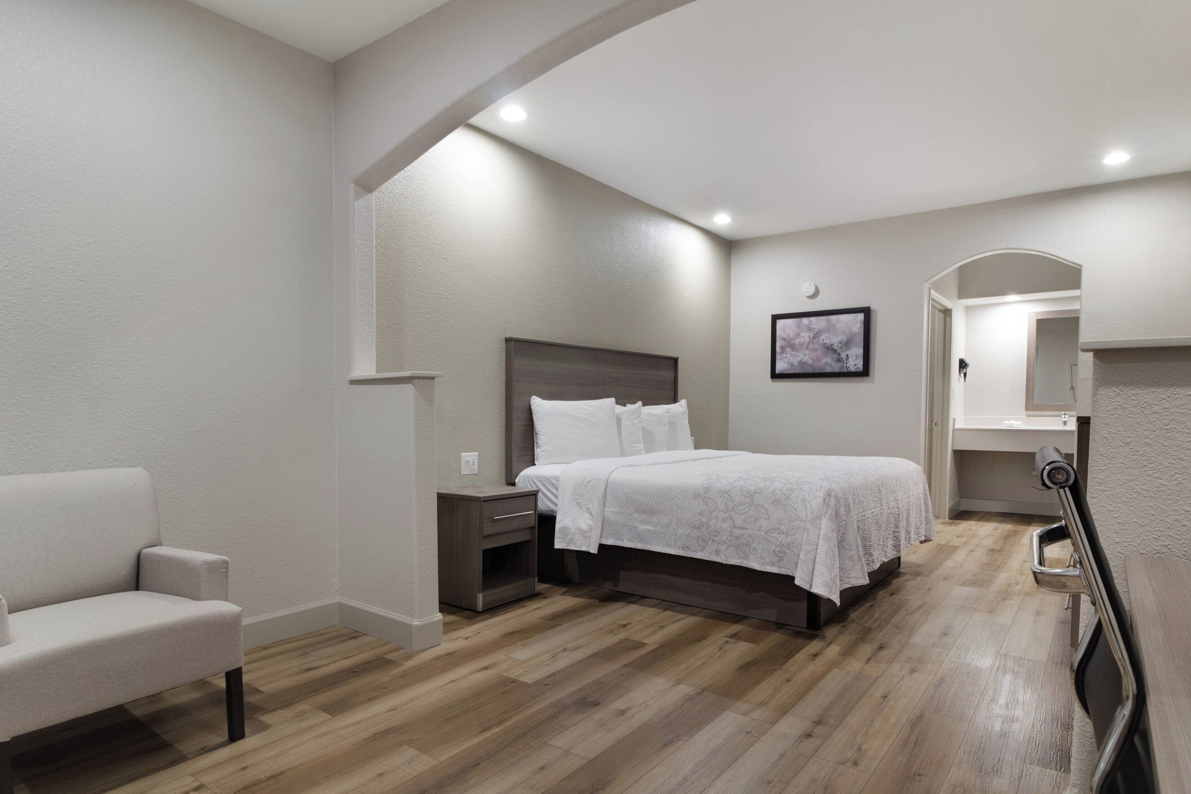 Un dormitorio con cama, mesita de noche, silla y un baño visible a través de un arco.