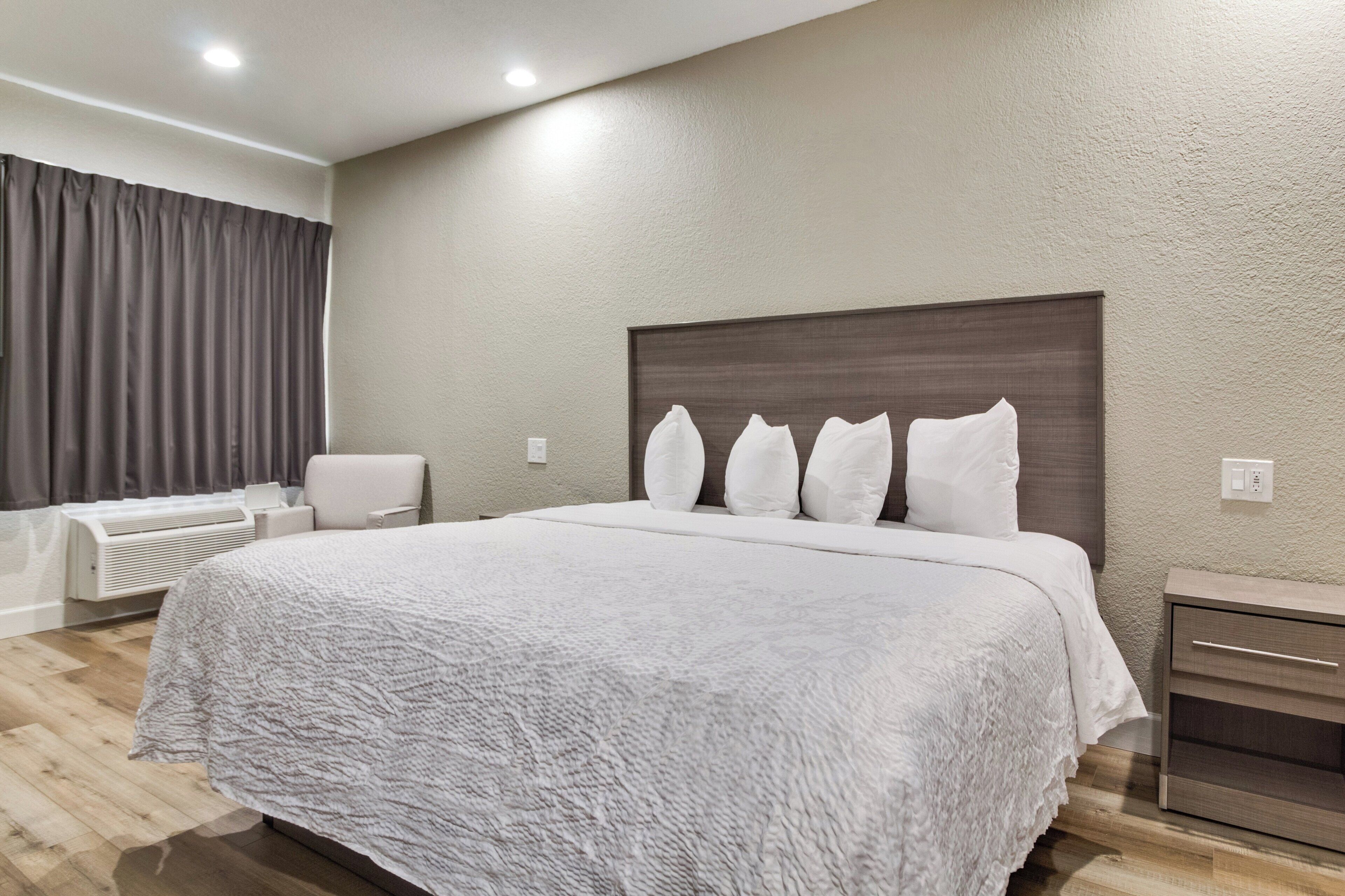 A modern hotel room with a large bed, a bedside table, a chair, and a built-in heater.