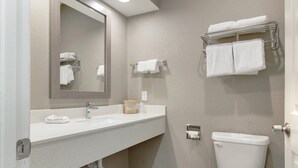 Un baño con lavamanos blanco, espejo e inodoro. Hay toallas en un perchero y un dispensador de jabón en el lavamanos.