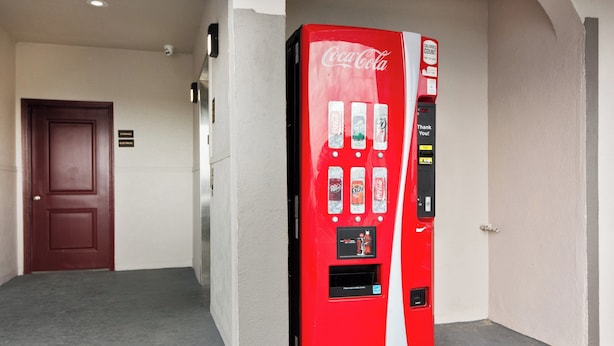 Una máquina expendedora roja de Coca-Cola en un pasillo con una puerta y un letrero que indica zonas de almacenamiento y eléctricas.