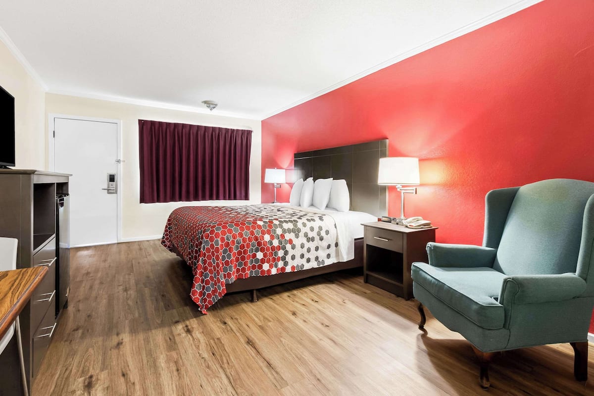 Quarto de hotel com cama, escrivaninha, cadeira, televisão e uma parede de destaque vermelha.
