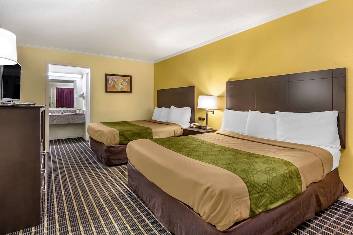 Quarto de hotel com duas camas, uma escrivaninha, uma cadeira, um espelho, uma luminária e um banheiro visível por uma porta aberta.