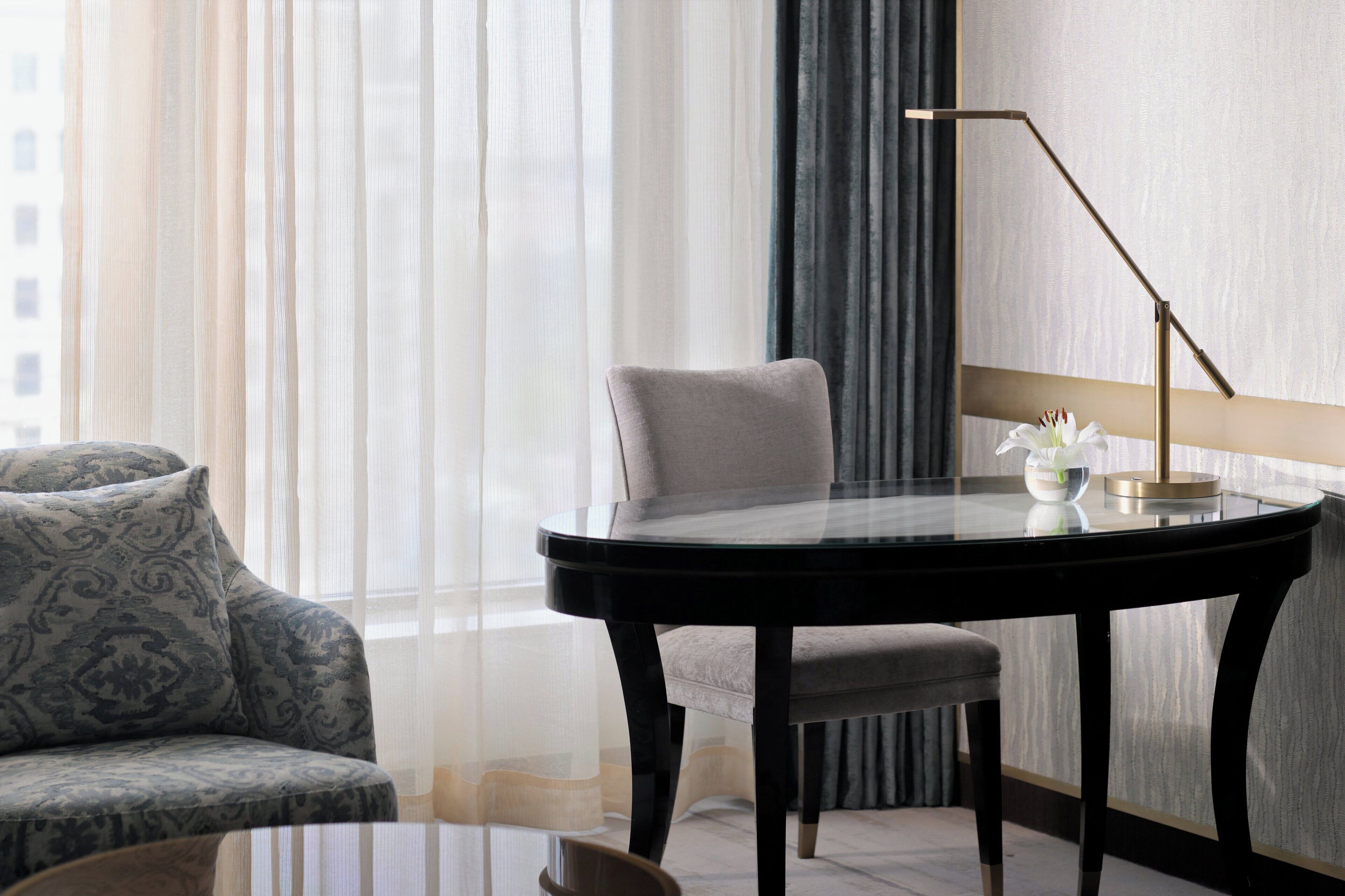 A modern hotel room with a glass-top table, a grey armchair, and a floor lamp.
