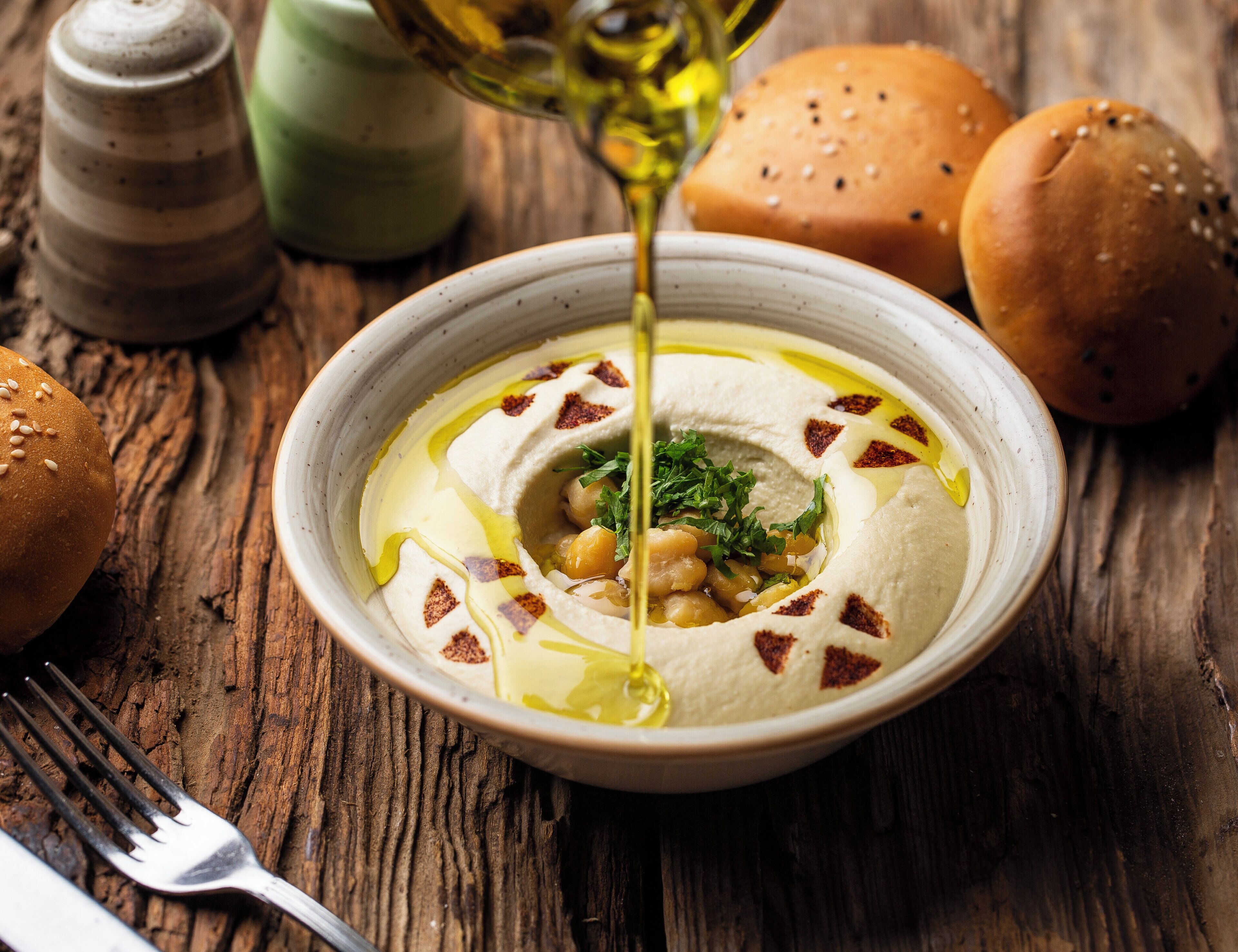 A bowl of hummus with olive oil being poured over it, surrounded by bread rolls and a fork on a wooden table.