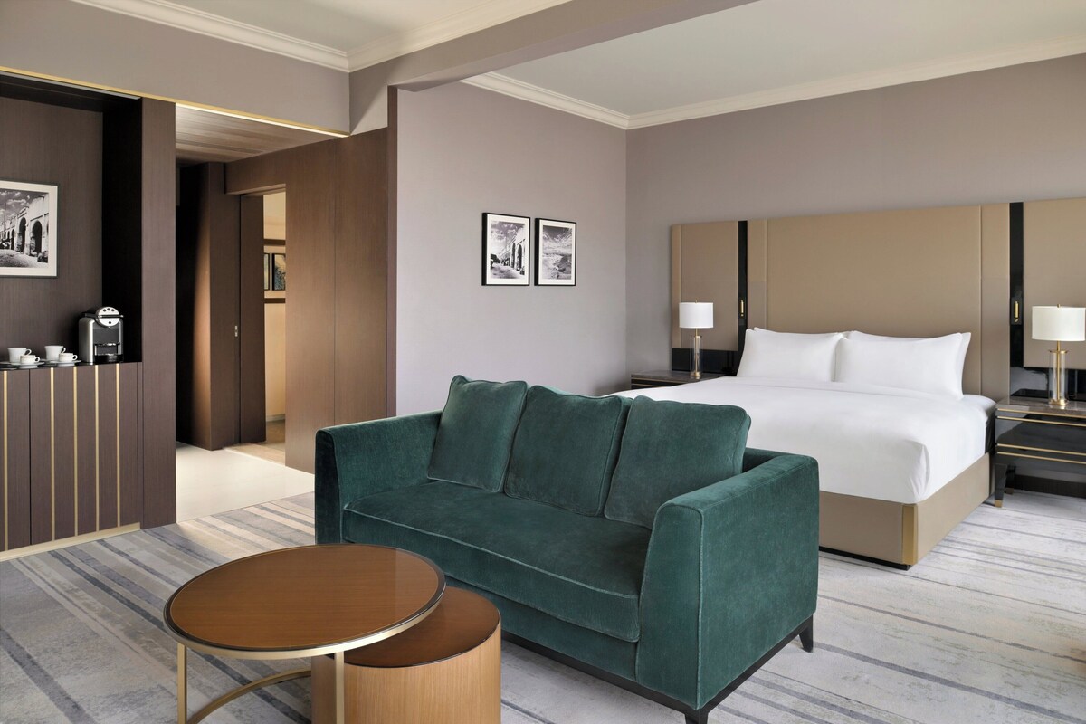 Ein Hotelzimmer mit einem grünen Sofa, einem runden Holztisch, einem Bett mit weißen Bettwäsche, einem Nachttisch mit Lampe und einer Kaffeemaschine.