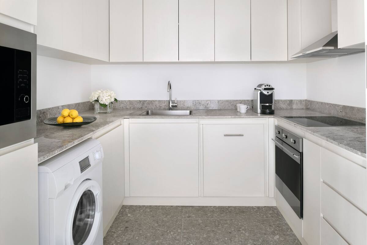 Cocina compacta con armarios blancos, lavadora integrada, cafetera y fregadero.