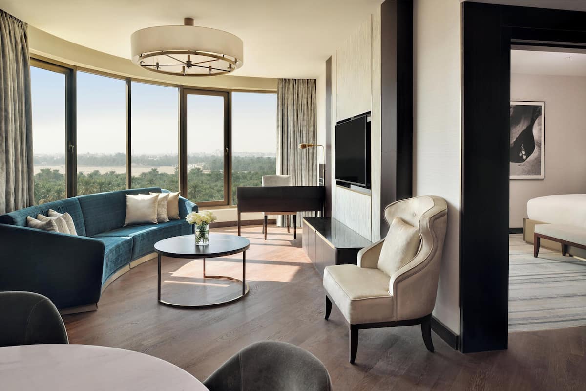 Habitación de hotel moderna con un ventanal amplio que ofrece vistas a la ciudad, un sofá, una silla, una mesita y un televisor de pantalla plana.