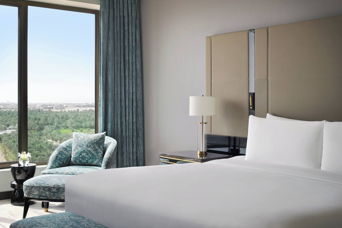 Habitación de hotel con una cama grande, una mesita de noche, una lámpara, una silla y vistas a la ciudad a través de amplios ventanales.
