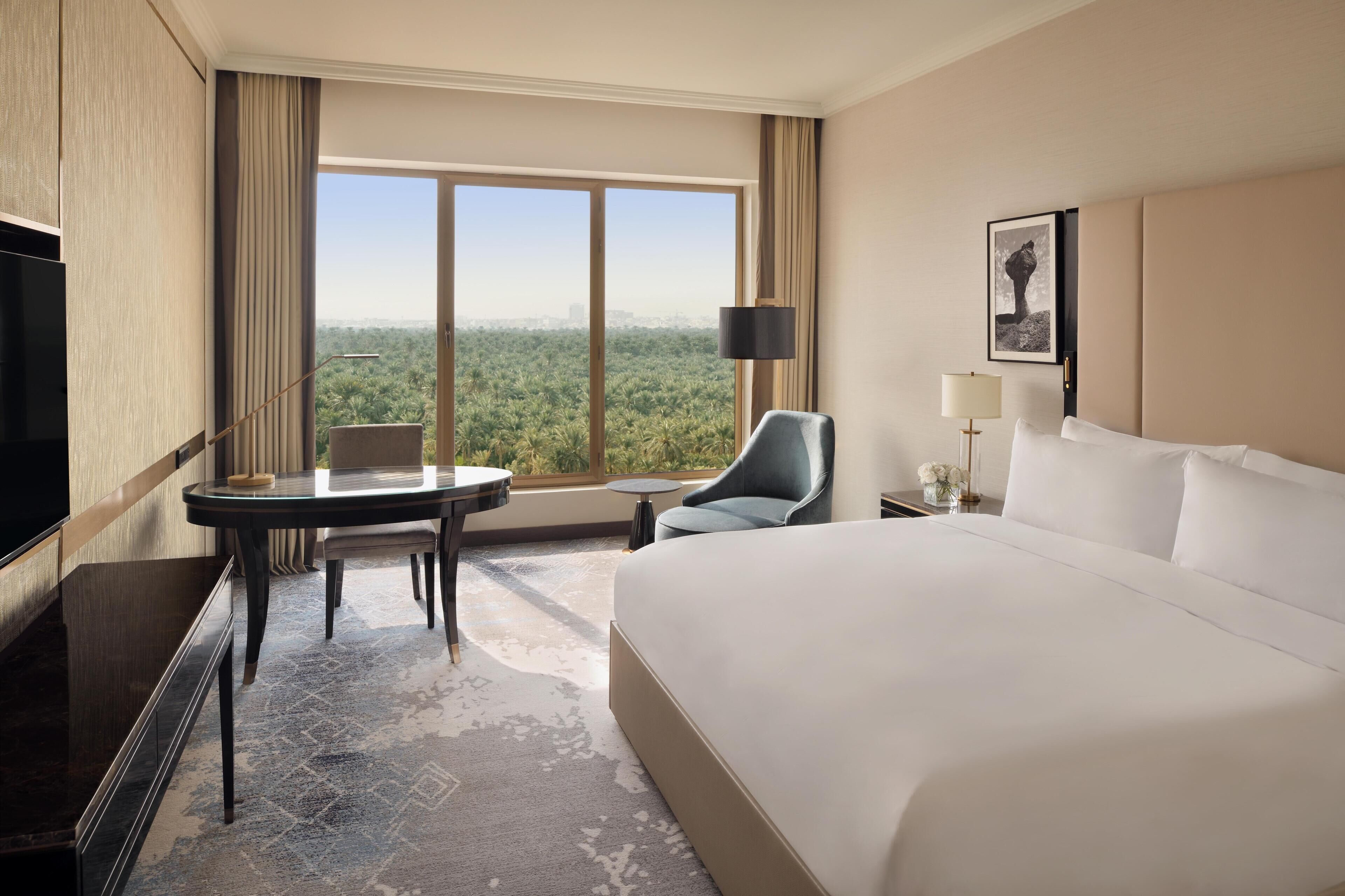 A hotel room with a large bed, a desk with a chair, a small table, a TV, a lamp, and a view of a cityscape through a large window.