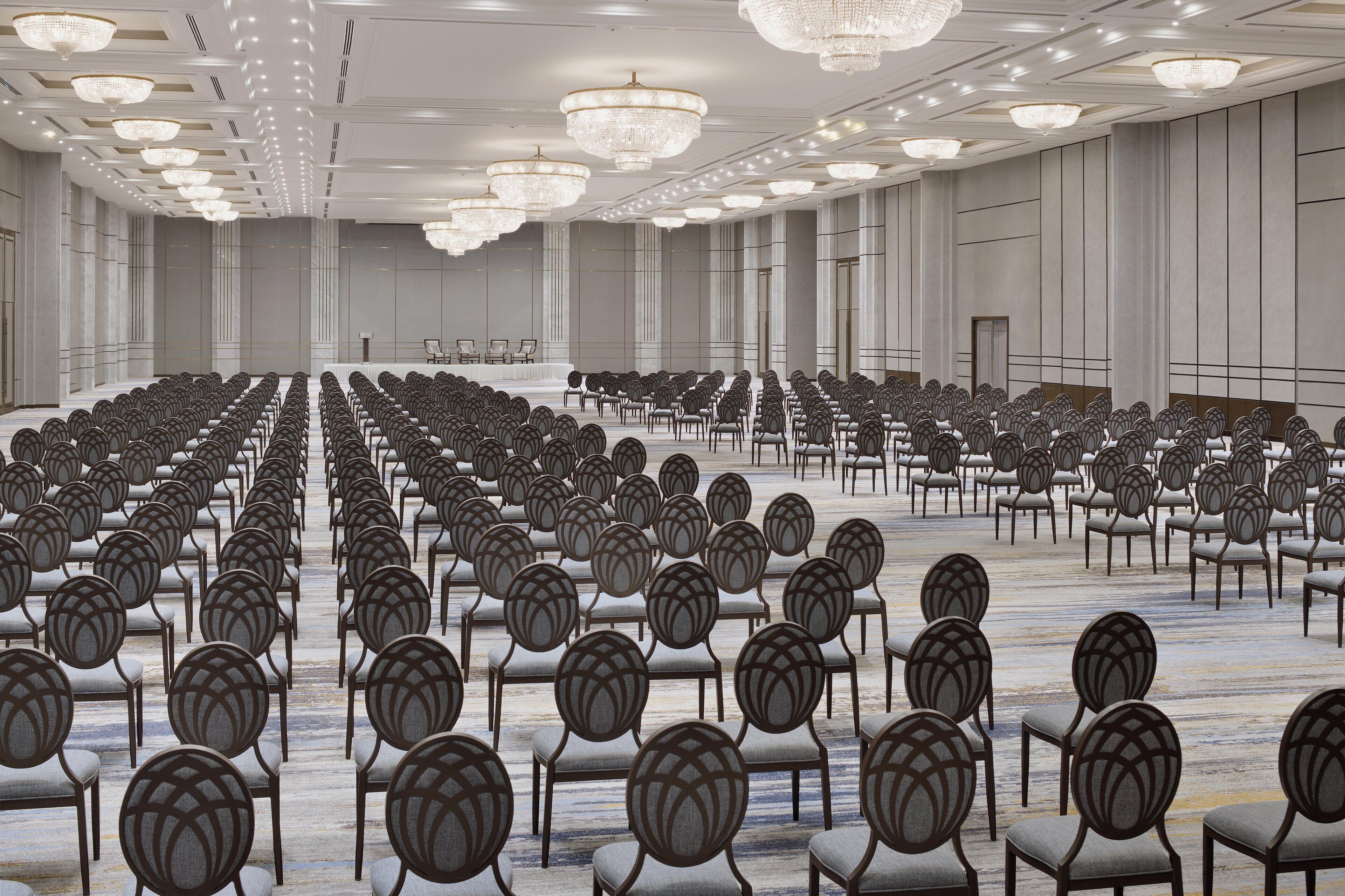 A large, well-lit conference room with rows of chairs facing a stage, featuring a modern chandelier and a neutral color scheme.