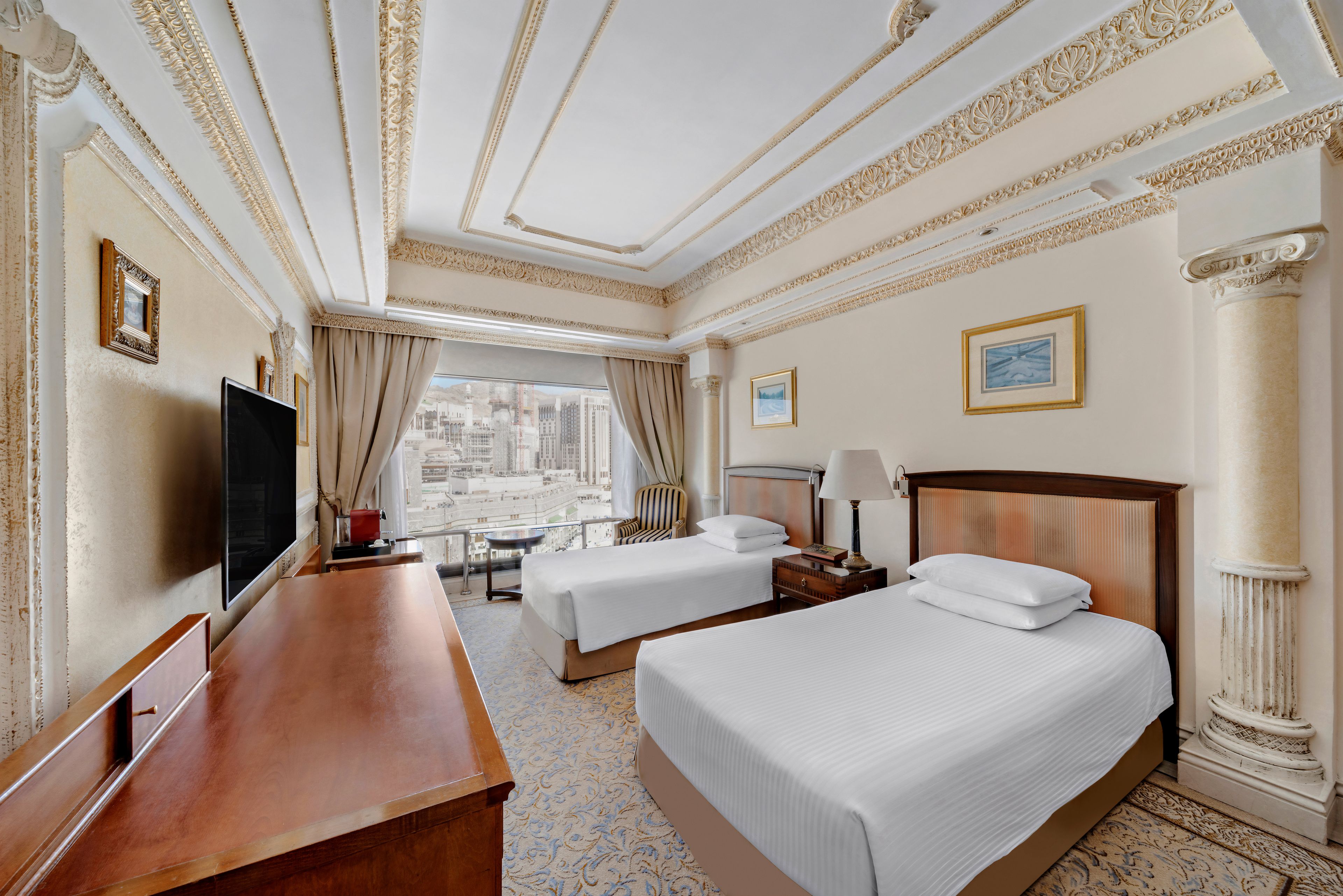 A hotel room with two beds, a desk, a TV, and a balcony with a city view.