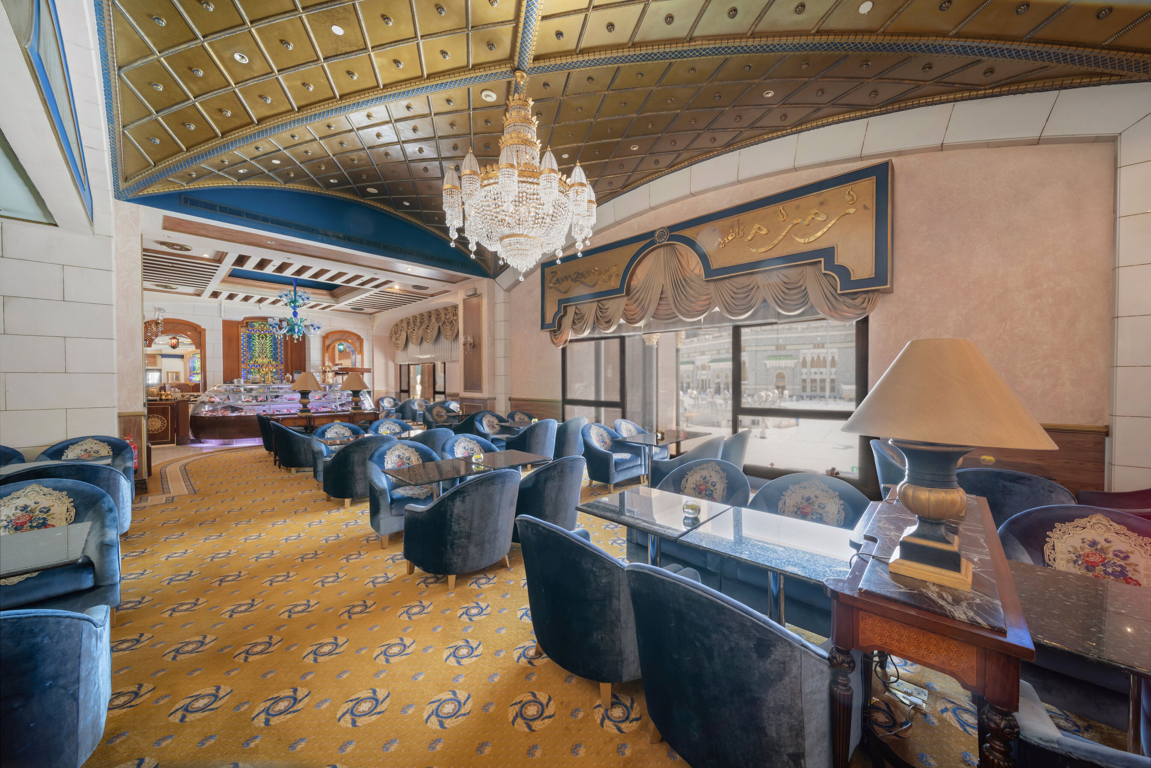 A spacious hotel lobby with a patterned carpet, blue upholstered seating, a chandelier, and a large window offering a view of the outside.