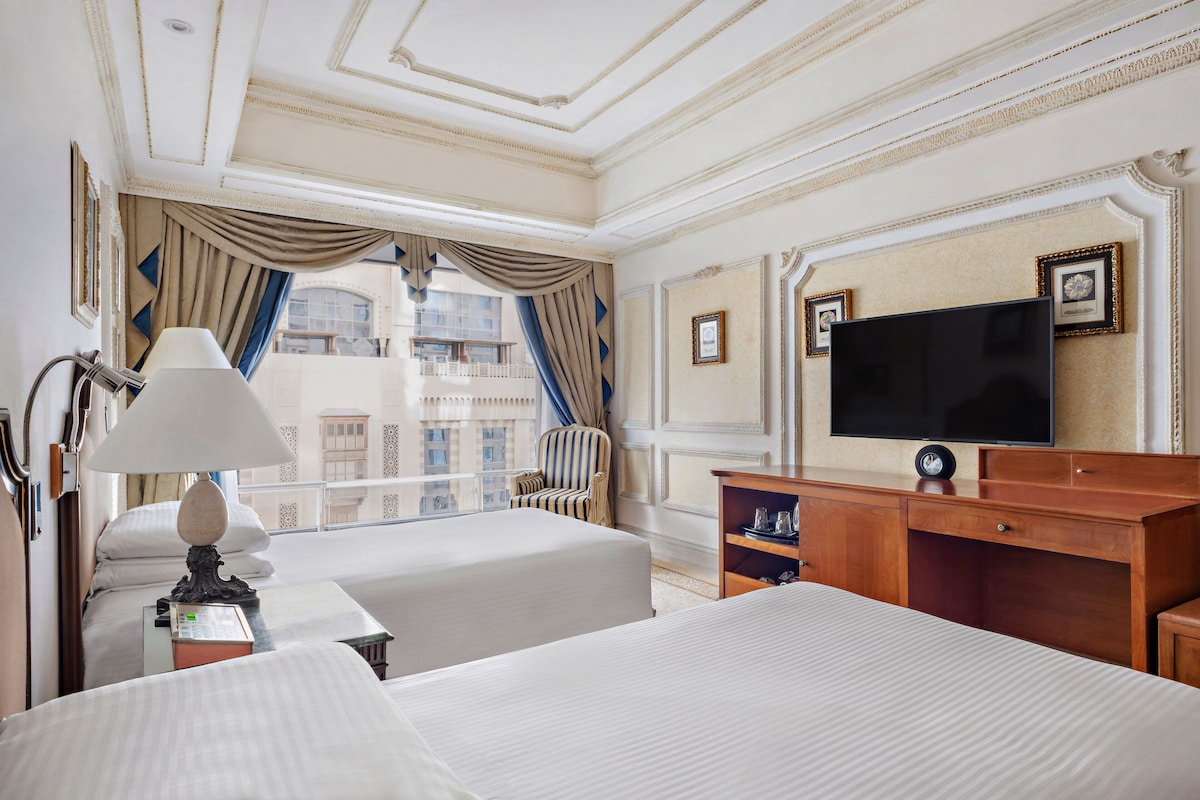 A hotel room with two beds, a television, a desk, and a view of the city.