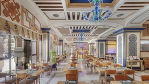 Un vaste espace de restauration d’hôtel, doté de nombreuses tables et chaises, d’un lustre et de panneaux décoratifs au plafond.